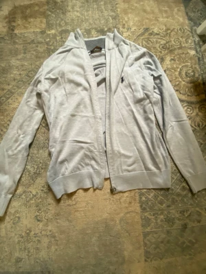 Ljusgrå cardigan från Polo Ralph Lauren - Ljusgrå cardigan performance från Polo Ralph Lauren med dragkedja framt tent och klassisk broderad logga rust på bröstet. Tröjan har ribbade muddar och är perfekt för lager-på-l radiostil. Enkel och stilren design som passar till det mesta sop.