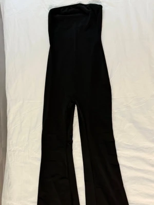 Svart tubjumpsuit med utsvängda ben - Säljer en svart, ärmlös tubjumpsuit med utsvängda ben. Jumpsuiten har en tight passform upptill och är gjord i ett stretchigt material som sitter snyggt på kroppen. Perfekt för dig som gillar en clean och stilren look.