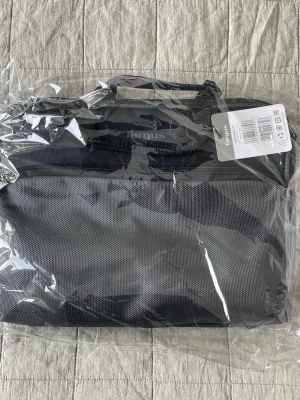 Svart datorväska från Targus - Snygg svart datorväska från Targus med praktiskt bärhandtag och dragkedja. Tillverkad i slitstarkt tygmaterial och har en framficka med dragkedja samt en genomskinlig ficka på baksidan för ID eller etikett. Perfekt för att bära din laptop.