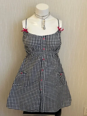 Rutig kortklänning med rosetter - Söt svartvit rutig kortklänning med tunna axelband och små rosa rosetter vid axlarna. Klänningen har knappar framtill och två fickor med rosa detaljer. Perfekt för sommarens alla tillfällen.