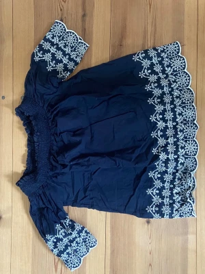 Blå offshoulder blus Abercrombie - Mörkblå offshoulder blus från Abercrombie & Fitch med vita broderade detaljer längs ärmar och nederkant. Blusen har korta ärmar med volangkant och smock upptill för en bekväm passform. Tillverkad i ett lätt tyg, perfekt för sommardagar.