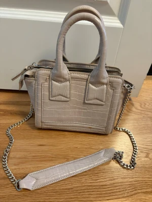 Beige croco-mönstrad handväska - Snygg beige axelremsväska från Zara  med croco-mönstrad yta. Väskan har både handtag och en axelrem i silverfärgad kedja med matchande axelparti. Perfekt storlek för det viktigaste och stängs med dragkedja.
