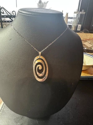 Silvrigt halsband med spiralhänge - Snyggt halsband med en tunn kedja i silverfärg och ett stort ovalt hänge med spiralformad design. Hänget har en cool, lite rå metallfinish . Perfekt för dig som gillar unika smycken med statement-känsla.