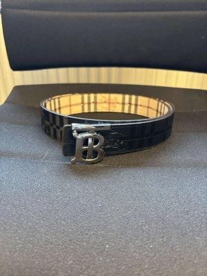 Svart rutigt Burberry bälte i skinn - Snyggt svart bälte från Burberry i äkta skinn med klassiskt rutigt mönster och en stor silvrig BB-logga som spänne. Perfekt accessoar för att lyfta din outfit med en exklusiv touch.