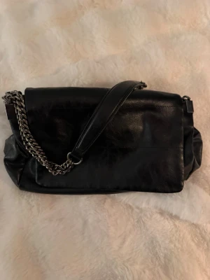 Zara - Svart axelväska i läder med kedja - Snygg svart axelväska i läder med både läderrem och silverfärgad kedja. Väskan har en klassisk rektangulär form och lockstängning. Perfekt för dig som gillar stilrena accessoarer med en tuff detalj.