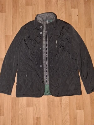 Svart och grön quiltad jacka med dragkedja - Stilren svart quiltad jacka med både knappar och dragkedja framtill. Jackan har hög krage, två framfickor och en bröstficka med dragkedja. Passar perfekt till kyligare dagar och har en klassisk look.