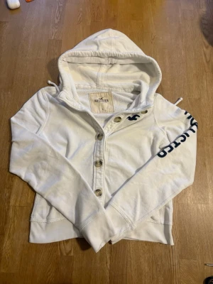 Hollister cardigan  - Vit hoodie från Hollister med knappstängning framtill och stor huva. Hollister-logga broderad i svart på ena ärmen och liten logga på bröstet. Tillverkad i mjukt bomullsmaterial, perfekt för en avslappnad och cool stil. Storlek l men sitter mer som S