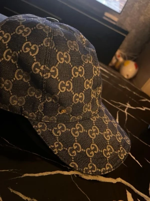  keps med GG-mönster - Snygg keps i mörkblå denim med det klassiska GG-monogrammet i beige över hela kepsen. Böjd skärm och justerbar passform. Perfekt för dig som vill ha en trendig accessoar med lyxig känsla och tydlig logga.