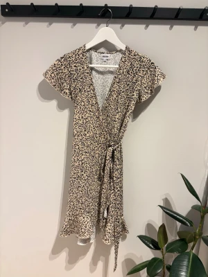 Leopardmönstrad omlottklänning - Kort leopardmönstrad omlottklänning med volangkant och knyt i midjan. Klänningen har korta ärmar och v-ringad hals. Beige botten med svarta leopardfläckar, tillverkad i mjukt och lätt tyg som passar perfekt för varmare dagar.