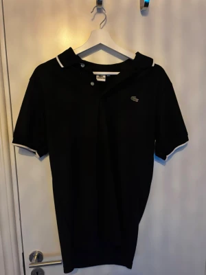 Svart polotopp från Lacoste - Säljer en svart polotopp från Lacoste med klassisk krage och vita detaljer på krage och ärmslut. Toppens material är bomull och den har korta ärmar. Den ikoniska Lacoste-loggan finns broderad på bröstet. Perfekt för dig som gillar stilrena plagg.