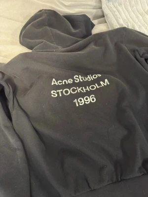 Svart hoodie från Acne Studios - Säljer en svart hoodie från Acne Studios med broderad text 'Acne Studios STOCKHOLM 1996' på ryggen. Klassisk passform med huva och ribbade muddar. Perfekt för dig som gillar stilrena och trendiga plagg. Materialet känns mjukt och skönt mot huden.