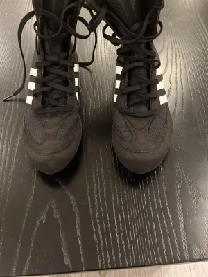 Adidas boxningsskor svart/vit – strl 38  - Säljer ett par stilrena boxningsskor från Adidas i svart med klassiska vita ränder. Lätta och bekväma skor som ger bra grepp och stabilitet – perfekta för boxning eller träning.  De ser smutsiga ut men det år hur modellen ser ut i verkligheten, kan skicka flera bilder privat om behov.                           • Storlek: 38 • Färg: Svart / vit • Skick: Bra skick • Höga för extra stöd vid ankeln  🥊 Perfekta för boxning, kampsport eller gym. 📦 Kan skickas inom 2 dagar. 💬 Skriv vid frågor!