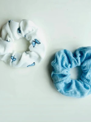 Två scrunchies i blått och vitt - Säljer två scrunchies, en vit med blå fjärilar i ribbat tyg och en ljusblå i mjukt, mönstrat tyg. Perfekta sopiga accessoarer för att piffa till din frisyr och soppa upp håret på ett snyggt sätt.