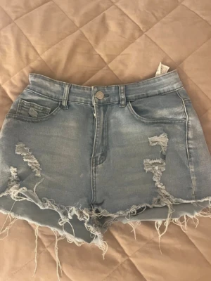 Ljusa jeansshorts med slitningar - Säljer ett par ljusblå jeansshorts med rå sopade kanter och slitna detaljer både fram och bak. Shortsen har fem fickor, bälteshällor och knappgylf. Perfekta för en avslappnad och trendig sommarlook.