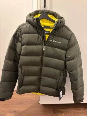  Sail Racing jacka  - Snygg olivgrön dunjacka från Sail Racing med gul insida och huva. Jackan har en puffig design, dragkedja framtill och logga på bröstet. Perfekt för kalla vinterdagar och riktigt varm tack vare dunfyllningen. Stilsäker och sportig look.