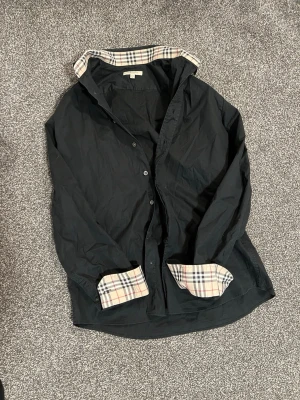 Burberry Skjorta – Svart med rutiga detaljer – M - Säljer en svart Burberry-skjorta i storlek M med rutiga detaljer som ger en klassisk och stilren look. Skjortan är i bra skick och behöver bara stryks. Passform bekväm med lite stretch tack vare 96% bomull och 4% elastan.  • Märke: Burberry • Storlek: M • Färg: Svart med rutiga detaljer • Material: 96% bomull, 4% elastan • Skick: Bra skick, behöver strykas Nypris: ca 4 400–5 300 kr Pris: 599 kr Kan skickas eller mötas upp.