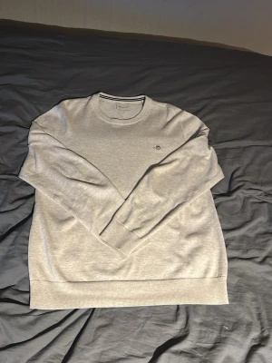 Beige stickad tröja från Gant - Snygg beige tröja från Gant med rund halsringning och diskret broderad logga på bröstet. Tröjan har långa ärmar och ribbade muddar vid ärmslut och nederkant. Perfekt för en clean och stilren look.