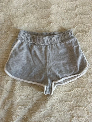 Grå sportiga shorts  - Säljer ett par grå shorts med vit kant och elastisk midja. De har en klassisk och enkel design som är perfekt för sommarens aktiviteter. Materialet är mjukt och bekvämt, storlek XS men större i storleken skulle jag säga. 