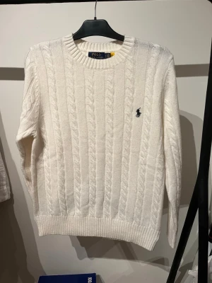 Vit kabelstickad tröja från Polo Ralph Lauren - Vit kabelstickad tröja från Polo Ralph Lauren med rund halsringning och broderad logotyp på bröstet. Tröjan har ribbade muddar vid ärmslut och nederkant, och är gjord i mjukt stickat material. Perfekt för att skapa en klassisk och stilren look.