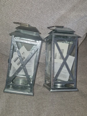 Ljuslyktor - Säljer två stilrena värmeljus lyktor i silverfärgad metall med glasväggar och handtag upptill. De har en klassisk fyrkantig form och X-detaljer på sidorna. Passar både inomhus och utomhus- att hänga eller ha ståendes. Aldrig använda men legat i ett skåp och är därför i ett behov av lite rengöring