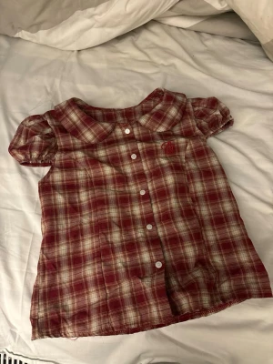 Röd- och vitrutigt blus med puffärm - Söt blus i röd- och vitrutigt mönster med puffiga korta ärmar och rund krage. Knäpps framtill med vita knappar. Tunt och lätt material, perfekt för varma dagar. Klassisk och avslappnad stil som passar till jeans eller kjol.