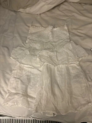 Vit somrig kortklänning med volang - Luftig och söt vit kortklänning med smock upptill och volangdetaljer. Klänningen är ärmlös och gjord i ett tunt, lätt tyg som passar perfekt för varma dagar. Enkel och stilren design som känns fräsch och trendig.