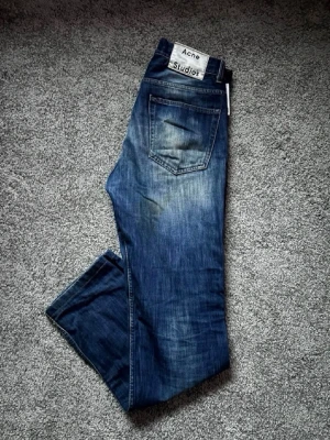 Blå raka jeans från Acne Studios - Säljer ett par klassiska blå jeans från Acne Studios med raka ben och femficksmodell. Jeansen har en snygg tvättad look med subtila slitningar och tydlig logga på bakfickan. Perfekta för en avslappnad och trendig stil. Materialet är denim i bomull.