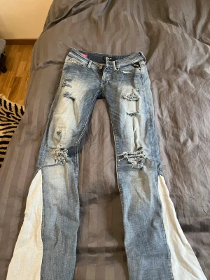 Replay jeans med slitningar  - Säljer ett par blå replay jeans med slitningar och hål på låren och knäna. Jeansen har unika vita tygpaneler längs utsidan av benen och en klassisk femficksmodell. Perfekta för dig som gillar en edgy och trendig stil. I storlek 23