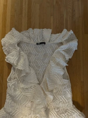 Vit volangblus från Zara - Säljer en vit blus från Zara med stora volanger längs kragen och framtill. Blusen är ärmlös och har ett strukturerat tyg i ett lätt, luftigt material. 