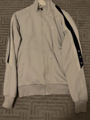  convenience Ljusgrå träningsdress G-Star Raw - Ljusgrå träningsdress från G-Star Raw med dragkedja framtill och ribbade muddar. Snygg svart och vit rand längs ena ärmen med RAW-tryck. Tillverkad i mjukt material, perfekt för sportiga outfits och chill dagar.