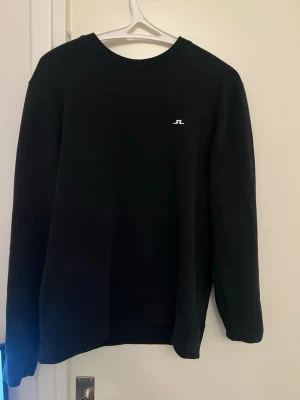 Svart sweatshirt från J.Lindeberg - Klassisk svart sweatshirt från J.Lindeberg med rund halsringning och diskret vitt JL-logotypbroderi på bröstet. Tröjan har långa ärmar och ribbade muddar vid ärmslut och nederkant. Perfekt för en clean och stilren look.
