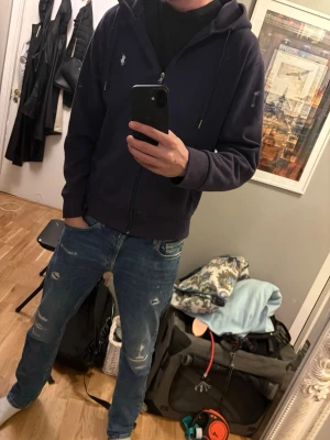 Mörkblå hoodie från Polo Ralph Lauren - Snygg mörkblå hoodie med dragkedja från Polo Ralph Lauren. Klassisk design med huva och dragsko, två fickor framtill och den ikoniska lilla loggan broderad på bröstet. Perfekt för en avslappnad och stilren look.