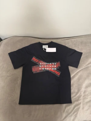 Svart t-shirt från Maison Margiela - Cool svart t-shirt från Maison Margiela med tryck i rött och vitt framtill. T-shirten har klassisk passform och rund halsringning. Perfekt för dig som gillar streetwear och unika designer.