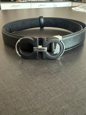 Svart skärp från Salvatore Ferragamo - Stilrent svart skärp från Salvatore Ferragamo med ikoniskt dubbel-Gancini-spänne i svart och silver. Bältet är gjord av äkta läder och har en exklusiv känsla som lyfter vilken outfit som helst.