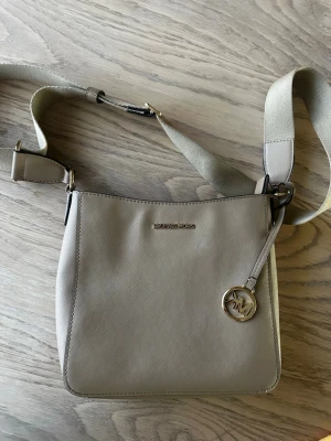 Beige axelväska från Michael Kors - Snygg beige axelväska från Michael Kors i stilren design med justerbar bred axelrem och guldfärgade detaljer. Väskan har en rund MK-berlock och en praktisk ytterficka på baksidan. Tillverkad i tåligt saffianoläder.