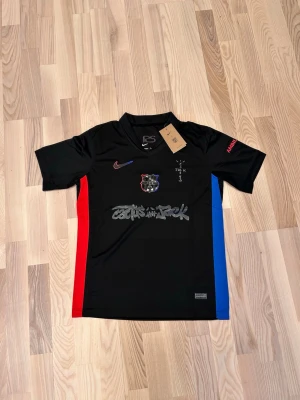Svart FC Barcelona x Cactus Jack tröja - Svart kortärmad fotbollströja från Nike i samarbete med FC Barcelona och Cactus Jack. Tröjan har röda och blå detaljer på sidorna, klubbmärke på bröstet och Cactus Jack-tryck framtill. Materialet är lätt och andas, perfekt för träning eller match.