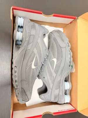 Nike Shox grå sneakers - Snygga Nike Shox sneakers i helgrå färg med vita detaljer och den klassiska Shox-dämpningen i hälen. Ovandelen är i mesh och syntet för en sportig look och bra ventilation. Perfekta för dig som gillar streetwear och vill sticka ut med en unik sula.