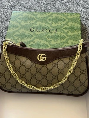Gucci GG Supreme axelremsväska - Snygg axelremsväska från Gucci i beige GG Supreme canvas med bruna läderdetaljer. Väskan har guldfärgad kedja och GG-logo framtill. Klassisk halvmåneformad modell med dragkedja upptill och exklusiv känsla. Perfekt för dig som gillar lyxiga accessoarer.