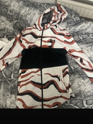 Under Armour hoodie med camomönster - Säljer en hoodie från Under Armour med coolt camomönster i vitt, svart och brunrött. Hoodien har hel dragkedja, fickor på sidorna och en stor huva. Perfekt för dig som vill sticka ut och gillar sportiga kläder.