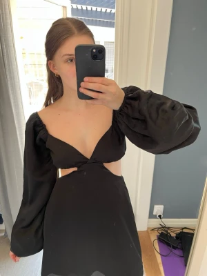 Svart kort klänning med puffärm - Trendig svart kort klänning med v-ringning, puffiga långärmar och cut out-detaljer i midjan. Klänningen har öppen rygg med en söt rosett🌷