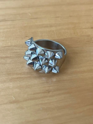 Silvrig ring med nitar från Edblad - Storlek 18.5 LCool och unik silvrig ring med flera små konformade nitar på ovansidan. Ringen har en bred design och är tillverkad i metall. Perfekt för dig som gillar en edgy och alternativ stil. 