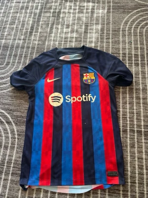 Barcelona matchtröja Nike blå/röd - Säljer en äkta FC Barcelona matchtröja från Nike med klassiska blå och röda vertikala ränder. Tröjan har klubbmärke och Nike-logga på bröstet samt Spotify som sponsor. Tillverkad i lätt och ventilerande material, perfekt för fotbollsträning eller match.