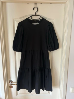 Svart klänning med puffärm från Zara - Svart klänning från Zara med puffärmar och rund halsringning. Klänningen har en öppen rygg med knyt och volangdetaljer i kjolen. 