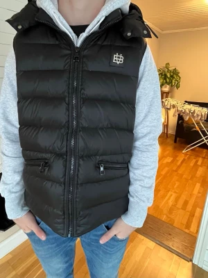 Svart dunväst från Bvaldi - Svart dunväst med quiltad design och två dragkedjeförsedda fickor framtill. Västen har en hög krage och logotyp på bröstet. Perfekt för lager-på-lager och håller dig varm under kyliga dagar. Stängs med dragkedja och har en stilren look.