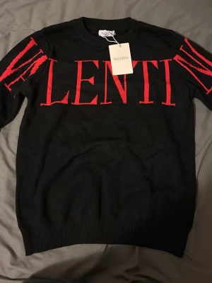 Svart stickad tröja från Valentino - Cool svart stickad tröja från Valentino med stora röda bokstäver över bröstet och ärmarna. Tröjan har rund halsringning och ribbstickade muddar. Perfekt för dig som vill ha en statement piece i garderoben.