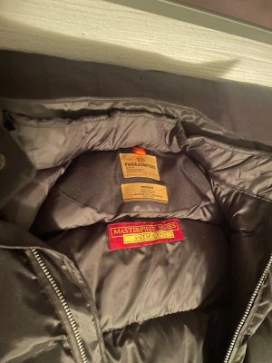 Svart dunväst från Parajumpers - Snygg svart dunväst från Parajumpers Masterpiece Series Anthology. Västen har quiltad design, dragkedja framtill och orange detaljer vid axlarna. Perfekt för lager-på-lager och håller dig varm med sin vadderade insida.