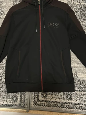 Svart hoodie från BOSS med röd dragkedja - Snygg svart hoodie från BOSS med röd dragkedja och diskret logga på bröstet. Hoodien har huva med dragsko och två fickor framtill. Perfekt för en stilren och sportig look.