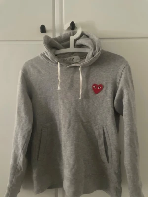 Grå hoodie från Comme des Garçons Play - Säljer en grå hoodie från Comme des Garçons Play med det ikoniska röda hjärtat med ögon broderat på bröstet. Hoodien har vit dragsko i huvan och två fickor framtill. Tillverkad i mjukt bomullsmaterial, perfekt för en avslappnad streetwear-look. ”OBS” tröjan sitter lite mindre i storlek 