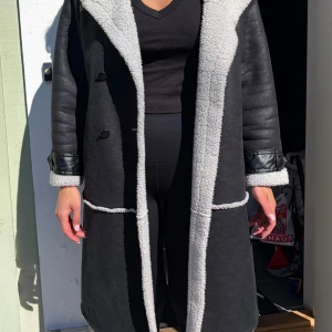 Svart lång fuskpäls-kappa M - Snygg svart lång kappa med vita fuskpälsdetaljer längs kanter, krage och ärmslut. Kappan har markerade sömmar och stängs med knappar framtill. Materialet ser ut som en mix av fuskpäls och fejkläder vilket ger en trendig look.lite större än M i modell .