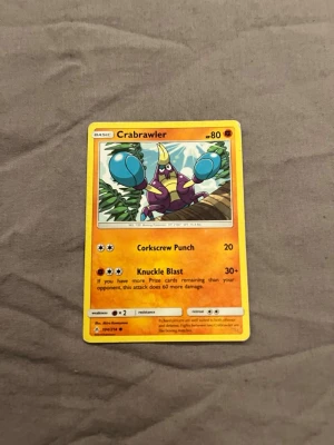 Pokémonkort Crabrawler - Säljer ett Pokémonkort med motivet Crabrawler. Kortet har en gul ram och färgstark illustration av Crabrawler med blå klor och lila kropp. Text och attacker står på engelska. Perfekt för samlare eller Pokémonfans.